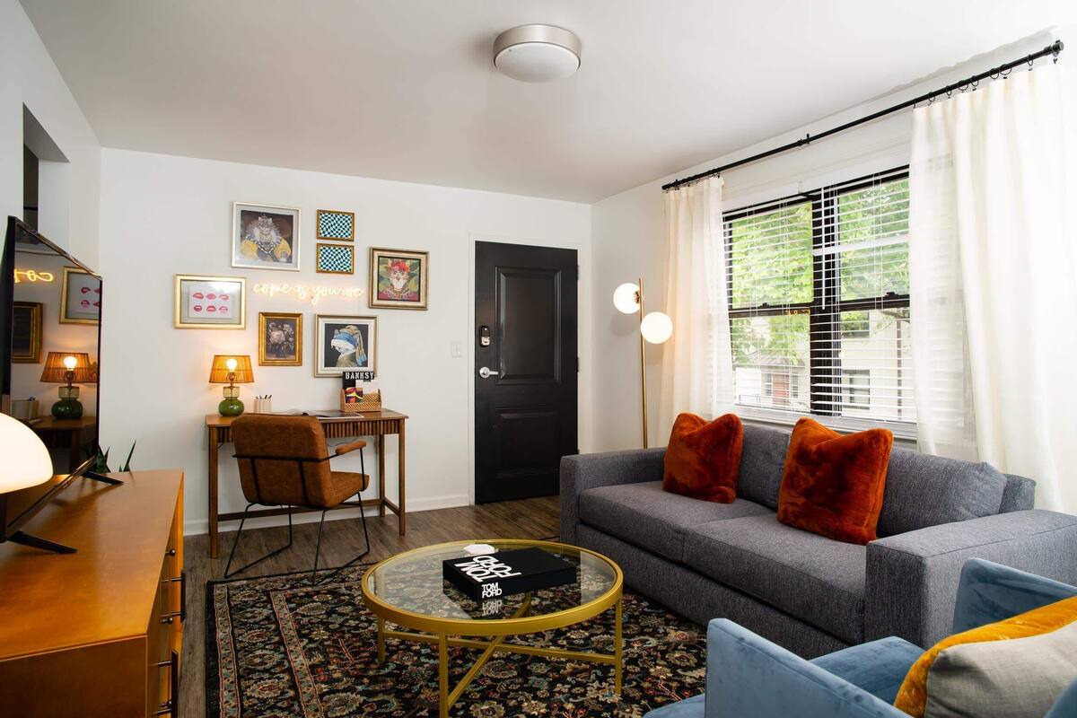 Een woonkamer met een bank, een salontafel, een stoel, een bureau en een boekenplank. Er is een deur en een raam met gordijnen.