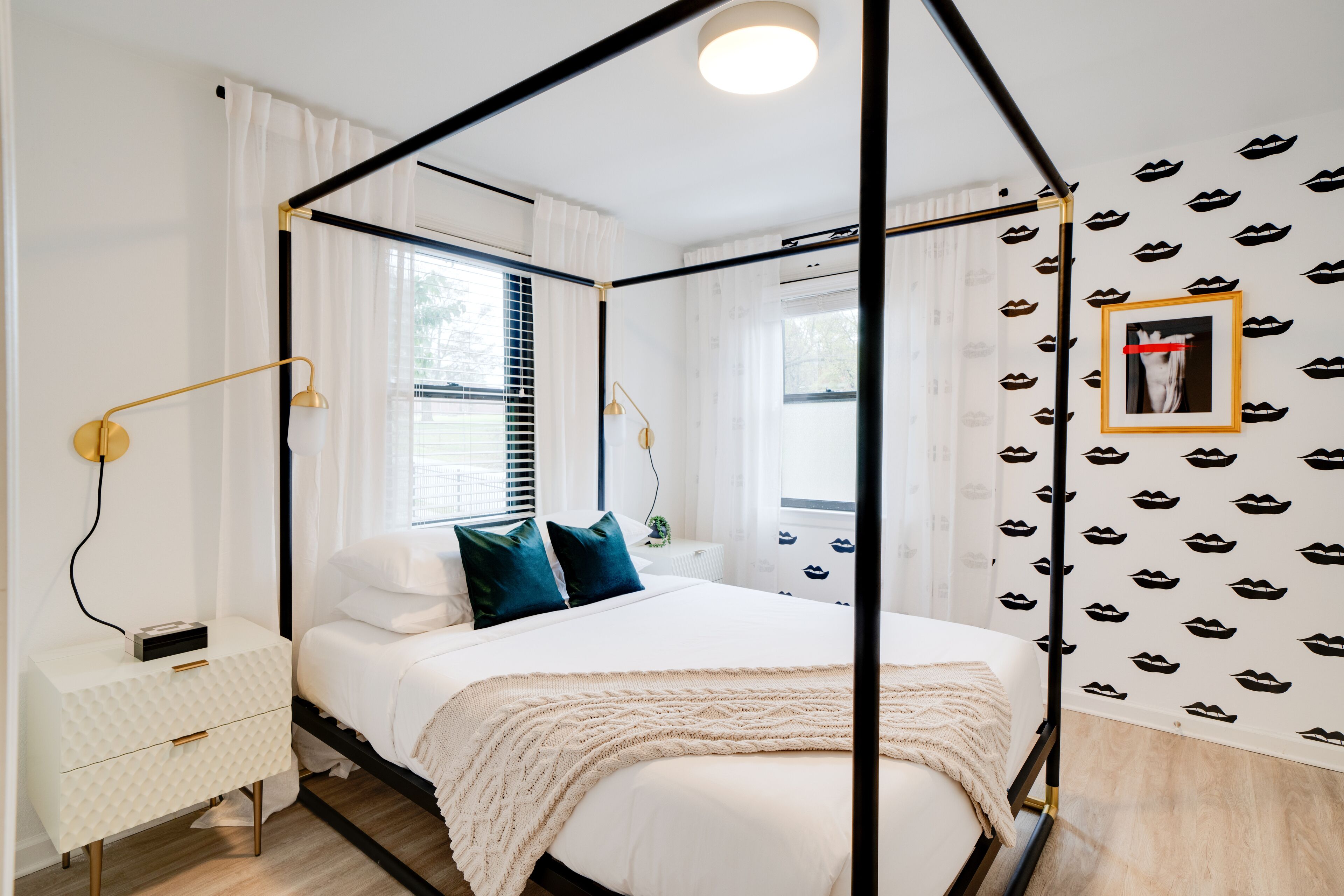 A four-poster bed with a white canopy, a bedside table with a lamp, and a wall with a pattern of lips.