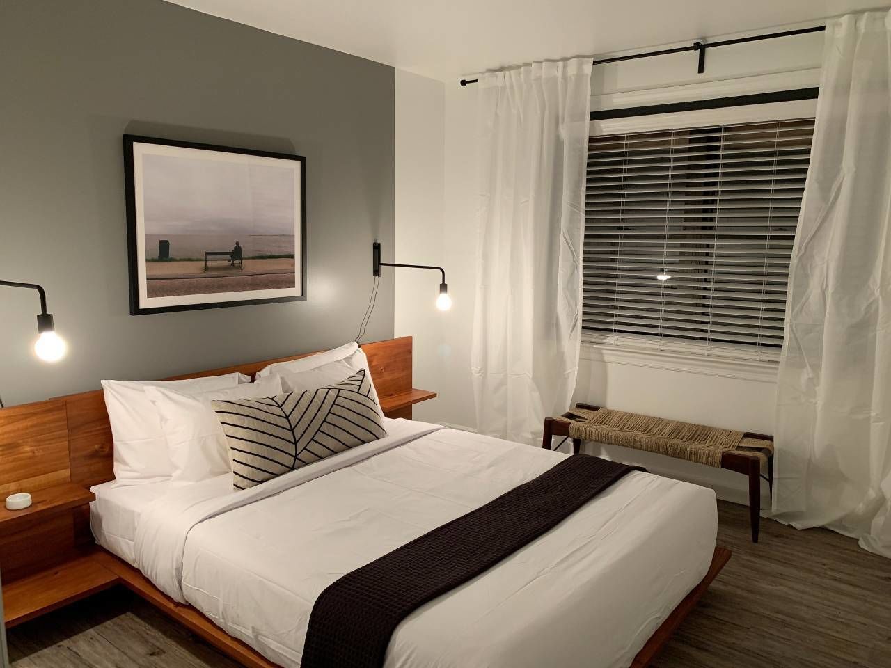 A neatly made bed with white linens, a wooden headboard, and a black throw blanket. There is a bench by the window and a framed picture on the wall.