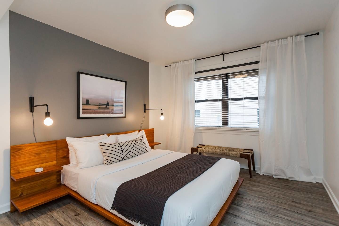 Een moderne slaapkamer met een houten hoofdbord, een groot bed met witte en gestreepte kussens, een nachtkastje, een bankje en een groot raam met zonwering.