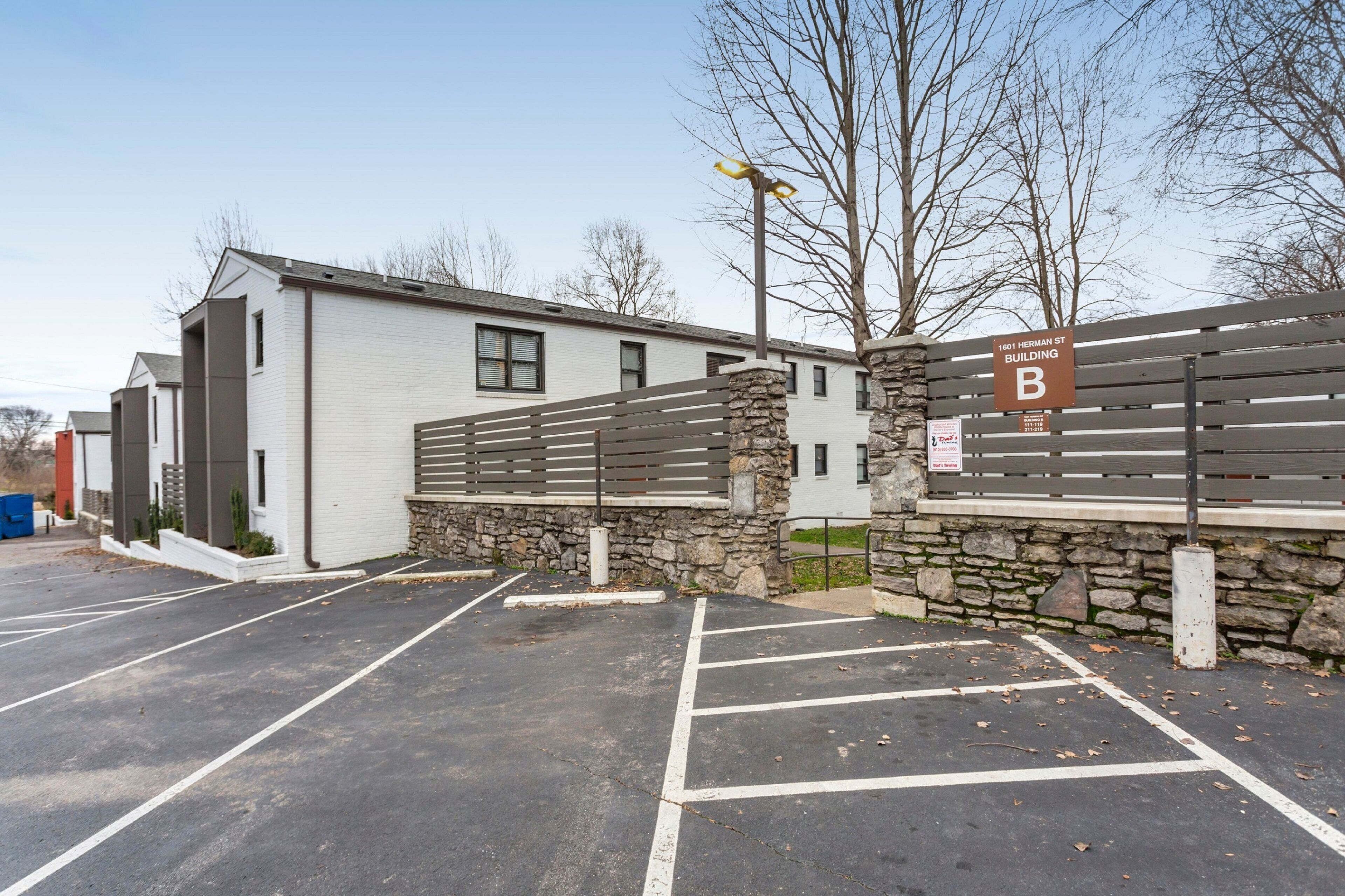 A parking lot with marked spaces, a building labeled "1601 Herman St Building B," and a stone wall with a metal fence.