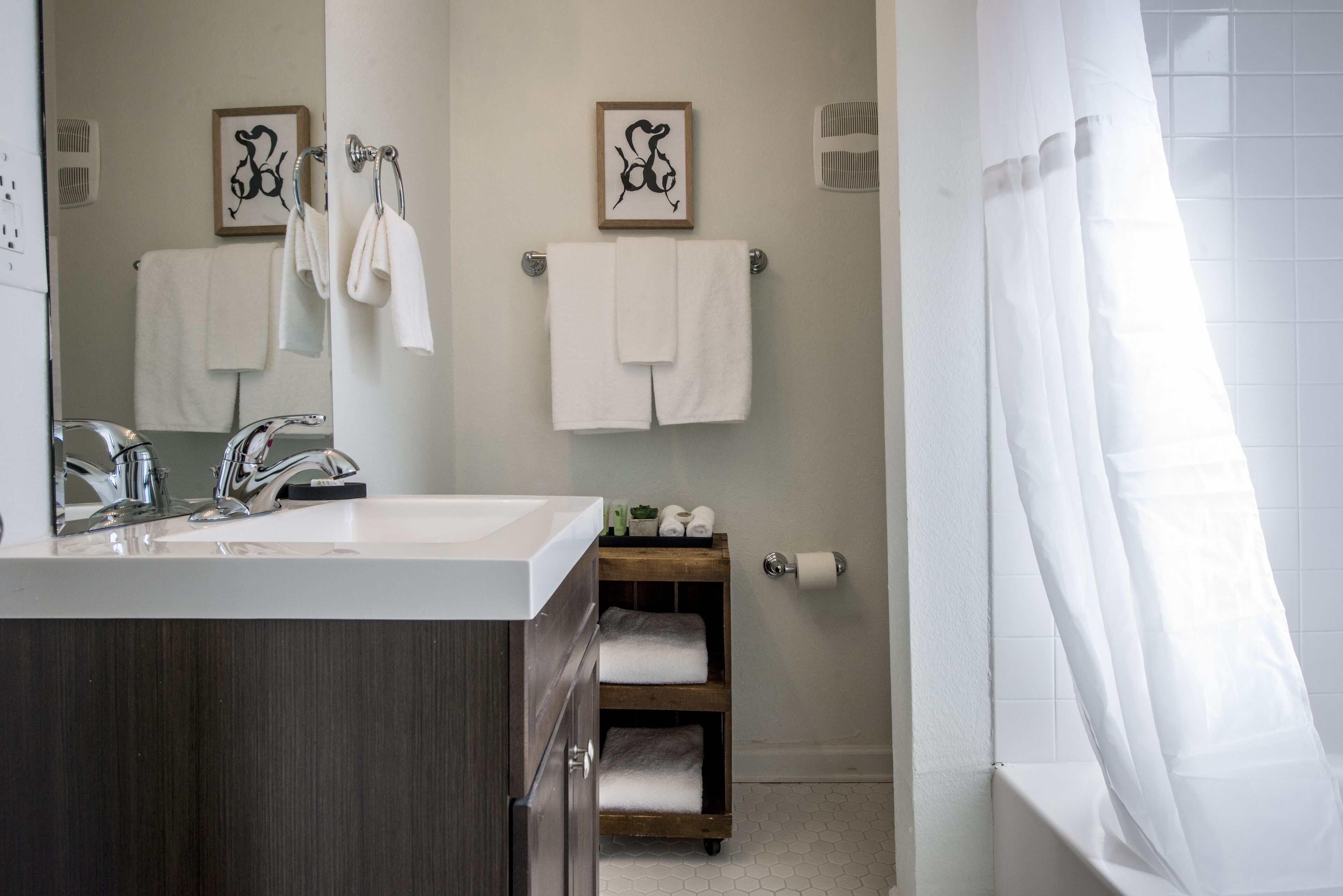 Une salle de bain moderne avec un lavabo blanc, deux porte-serviettes, une étagère en bois avec des serviettes et un rideau de douche.