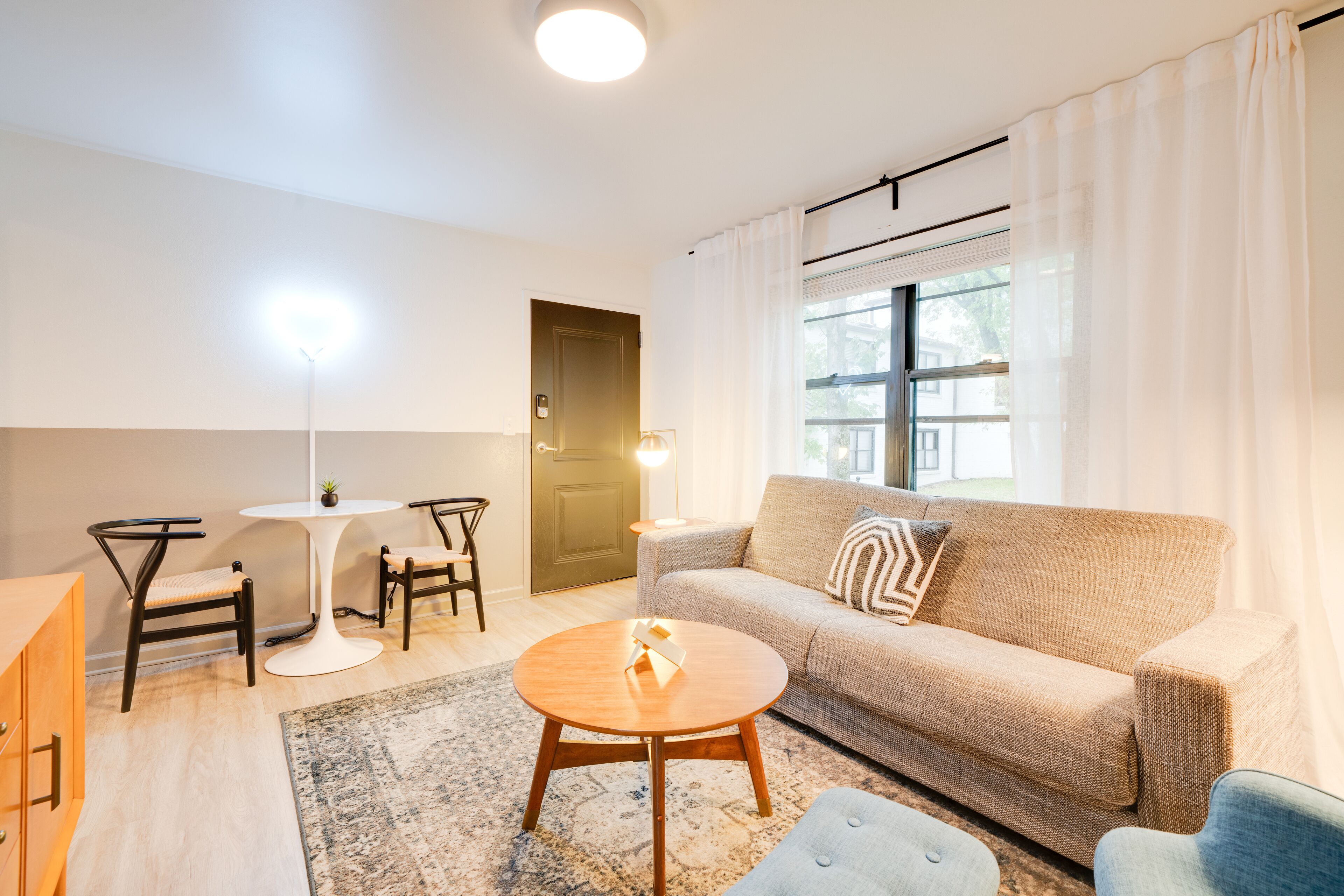 A living room with a sofa, two chairs, a round coffee table, a small table with a lamp, and a window with curtains.