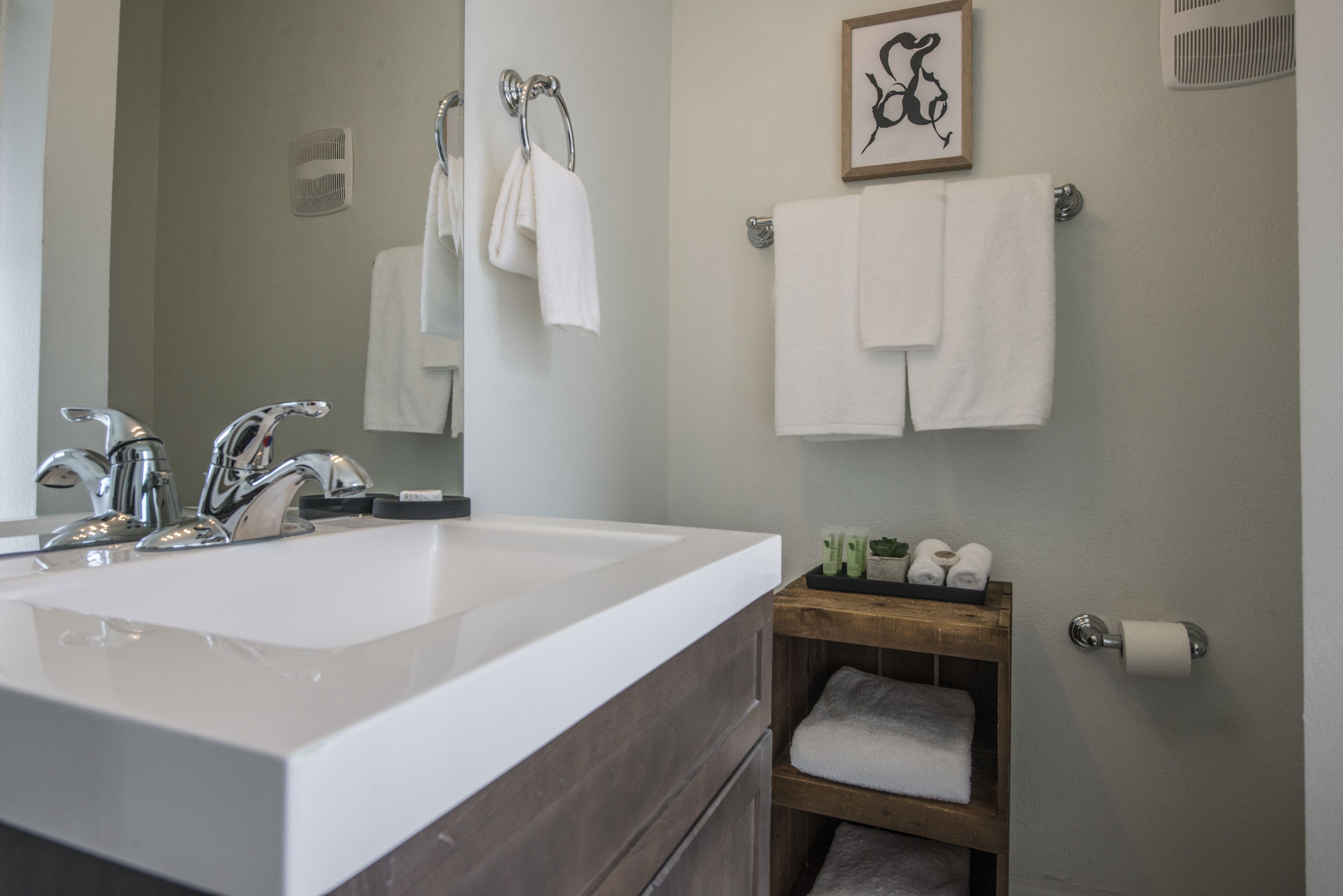 Un baño con un lavamanos grande de color blanco, dos grifos, un espejo y un estante de madera con toallas y artículos de aseo.