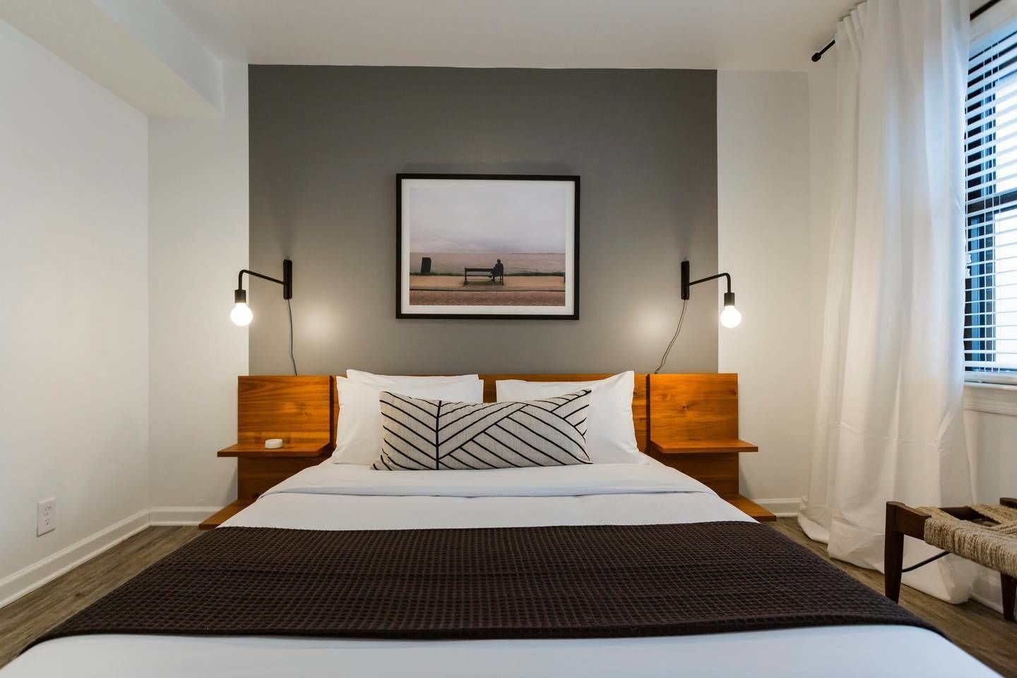 A modern bedroom with a large bed, two bedside tables, a wall-mounted picture, and a window with blinds.