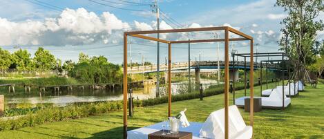 A serene outdoor setting with a bed and a canopy, a grassy area, a river, and a bridge in the background.