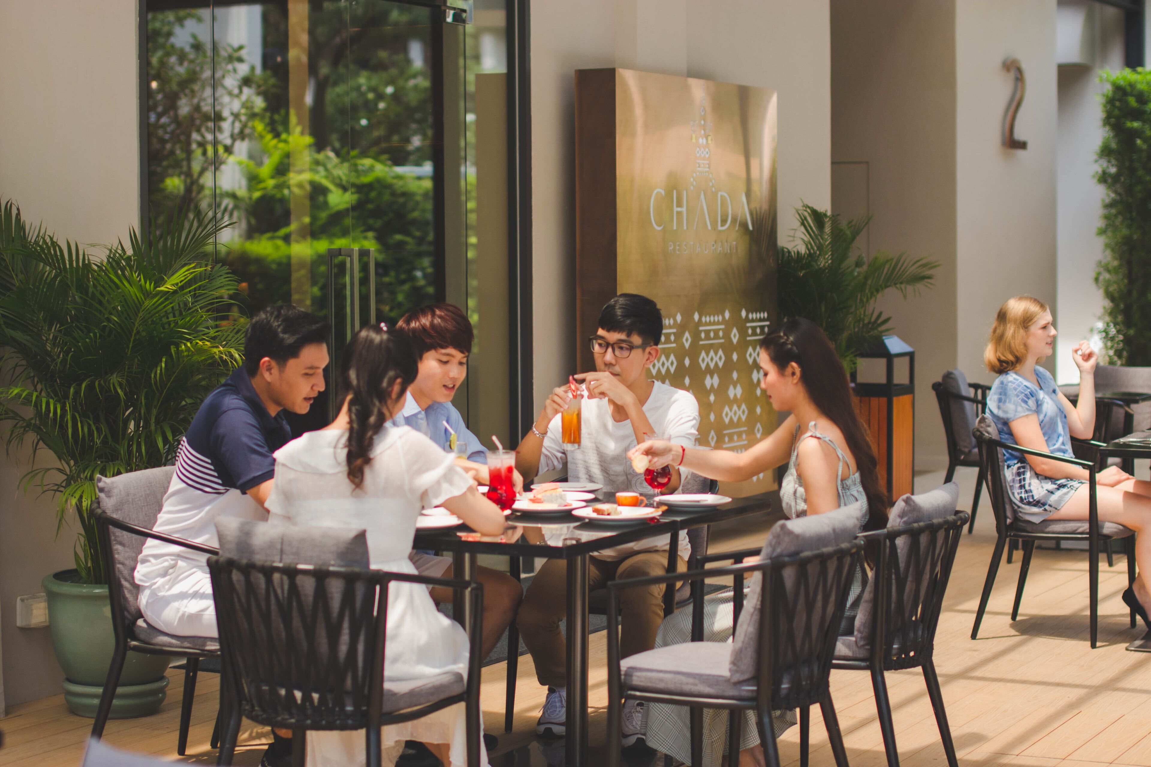 Un grupo de personas cenando en una mesa al aire libre con vistas a zonas verdes y un edificio con un letrero que dice "CHADA RESTAURANT".