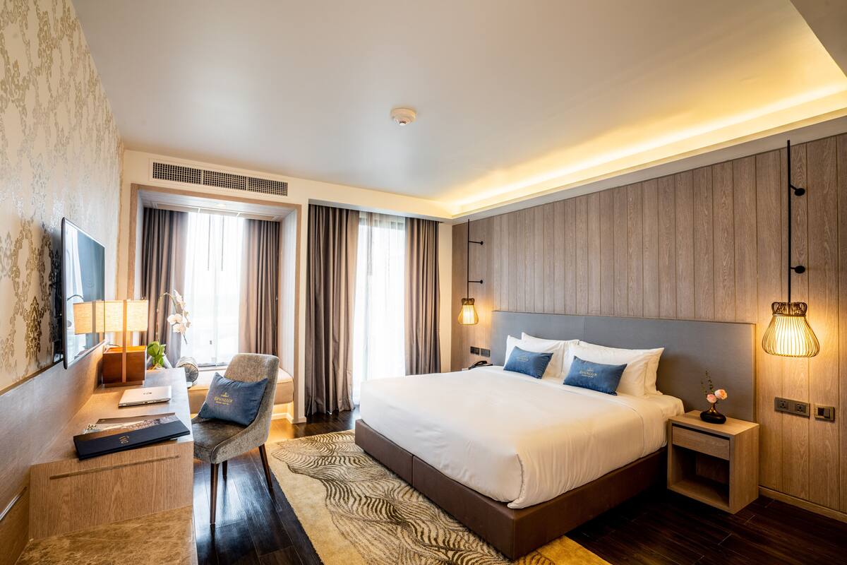 A modern hotel room with a large bed, a desk with a laptop, a chair, and a nightstand with a lamp.