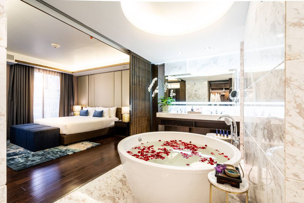 A modern hotel room with a large bed, a bathtub with rose petals, a vanity with a mirror, and a view of the city.