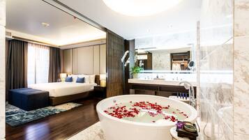 A modern hotel room with a large bed, a bathtub with rose petals, a vanity with a mirror, and a view of the city.