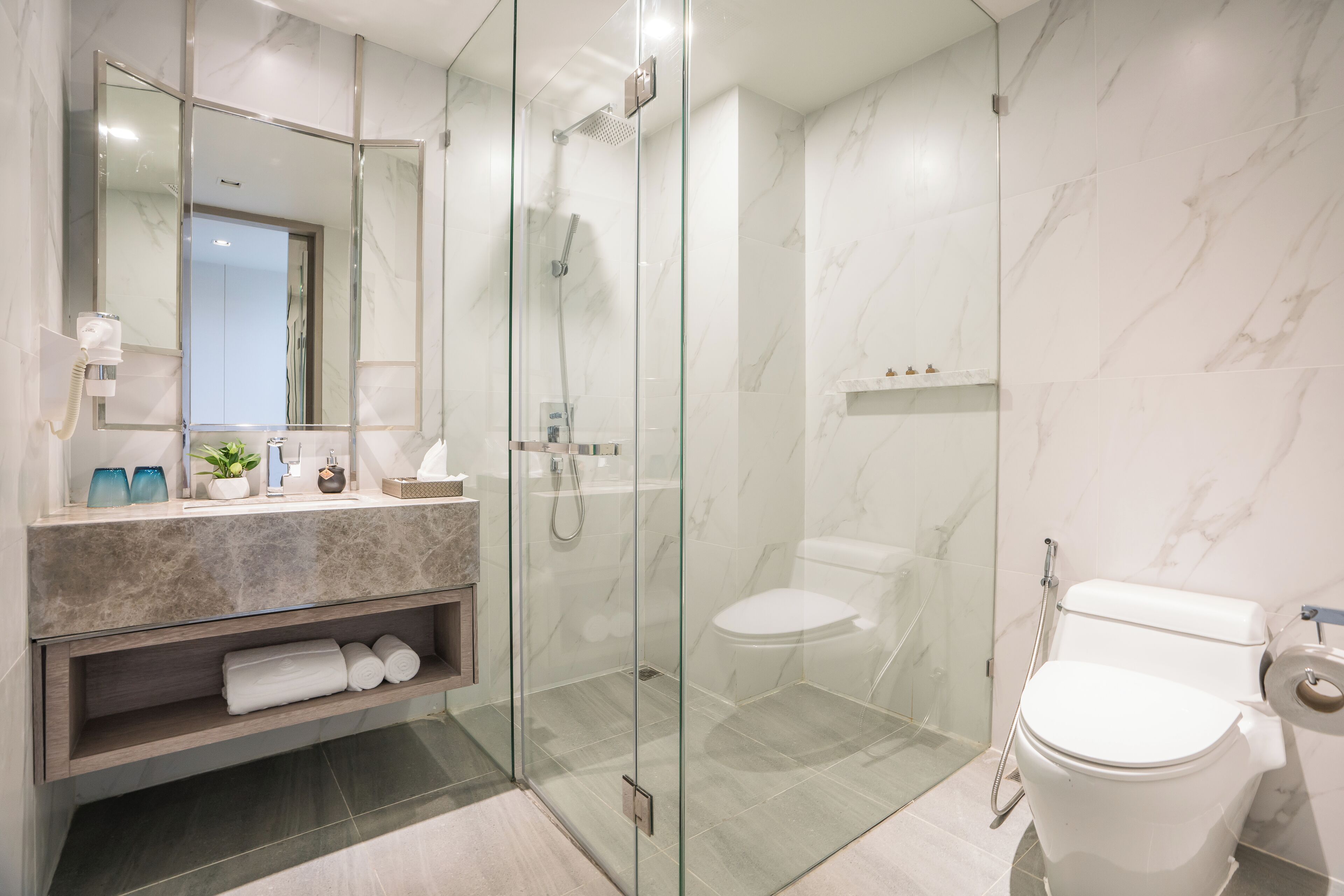 Un baño moderno con ducha envuelta en vidrio, pared con mosaicos de mármol, inodoro blanco y tocador con espejo y toallero.