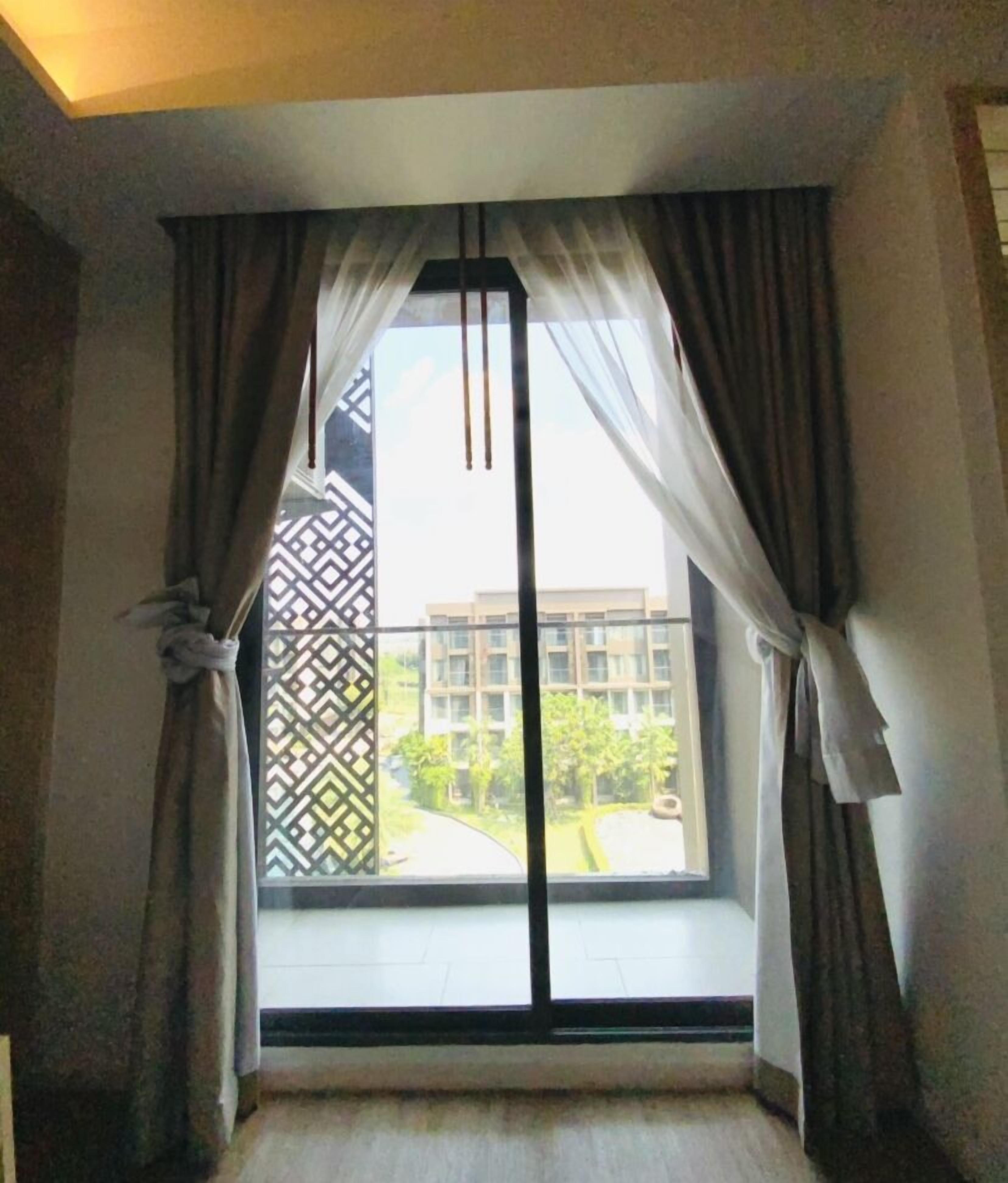 A room with a large window, curtains, and a view of a building and greenery outside.