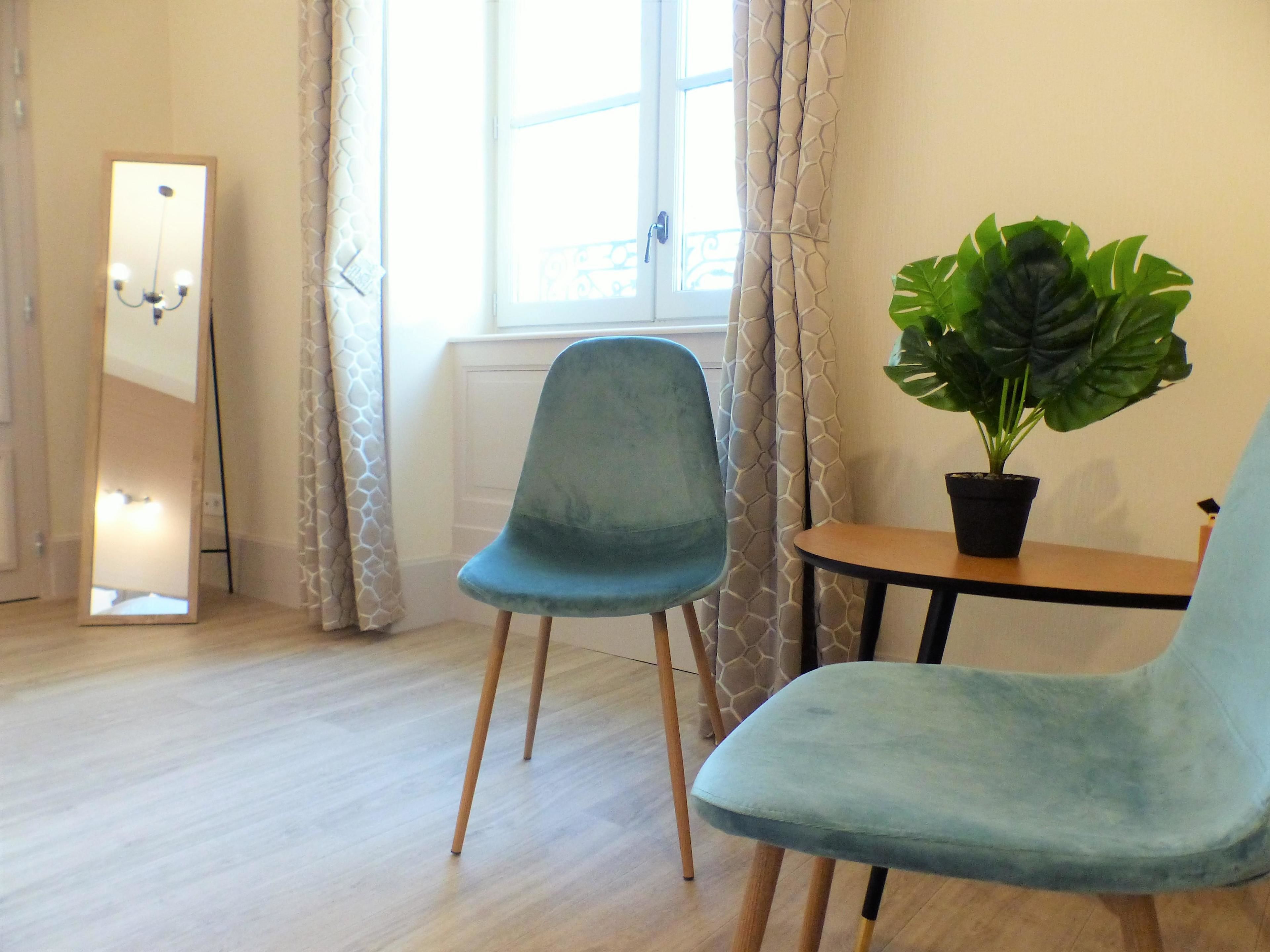 Une pièce avec deux chaises bleues, une petite table sur laquelle est posée une plante en pot, un miroir et une fenêtre avec des rideaux.