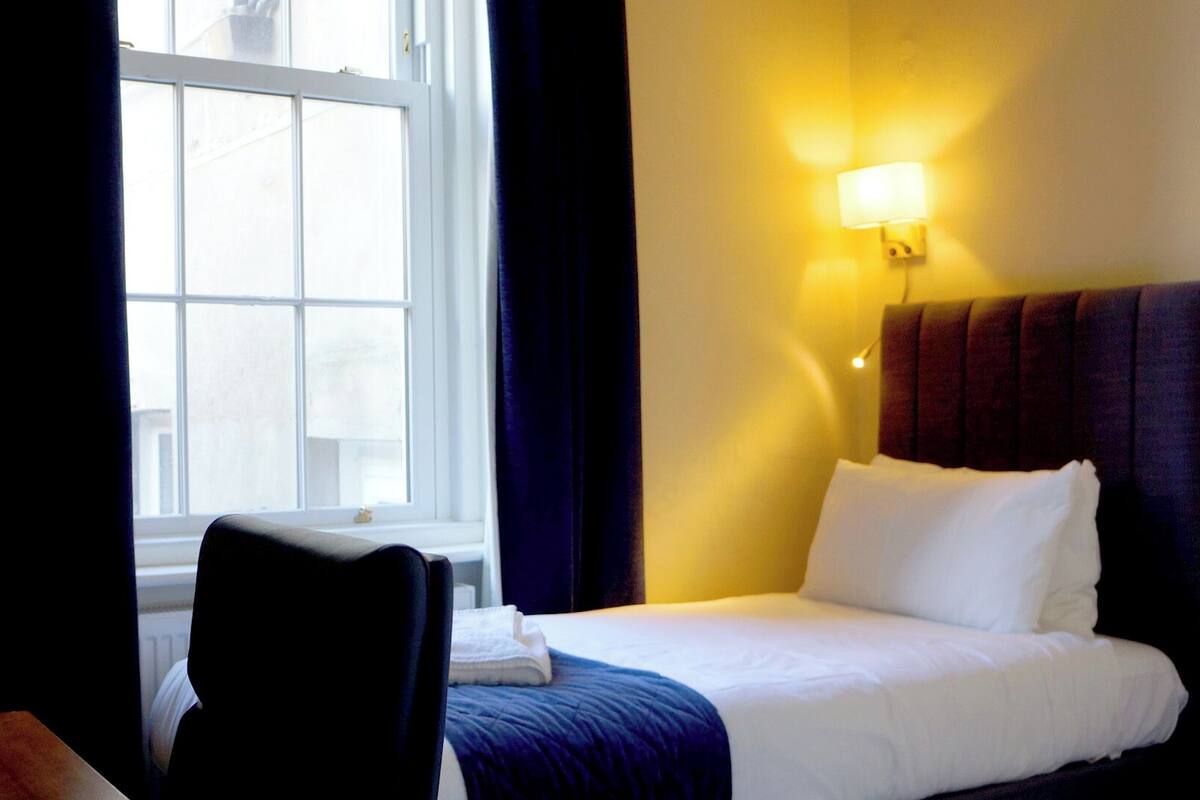 A hotel room with a bed, a chair, a window, and a wall-mounted light.