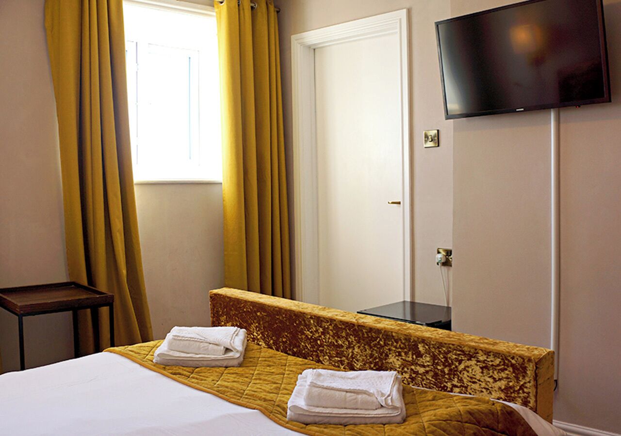 A hotel room with a bed, a bedside table, a TV mounted on the wall, and a window with curtains.