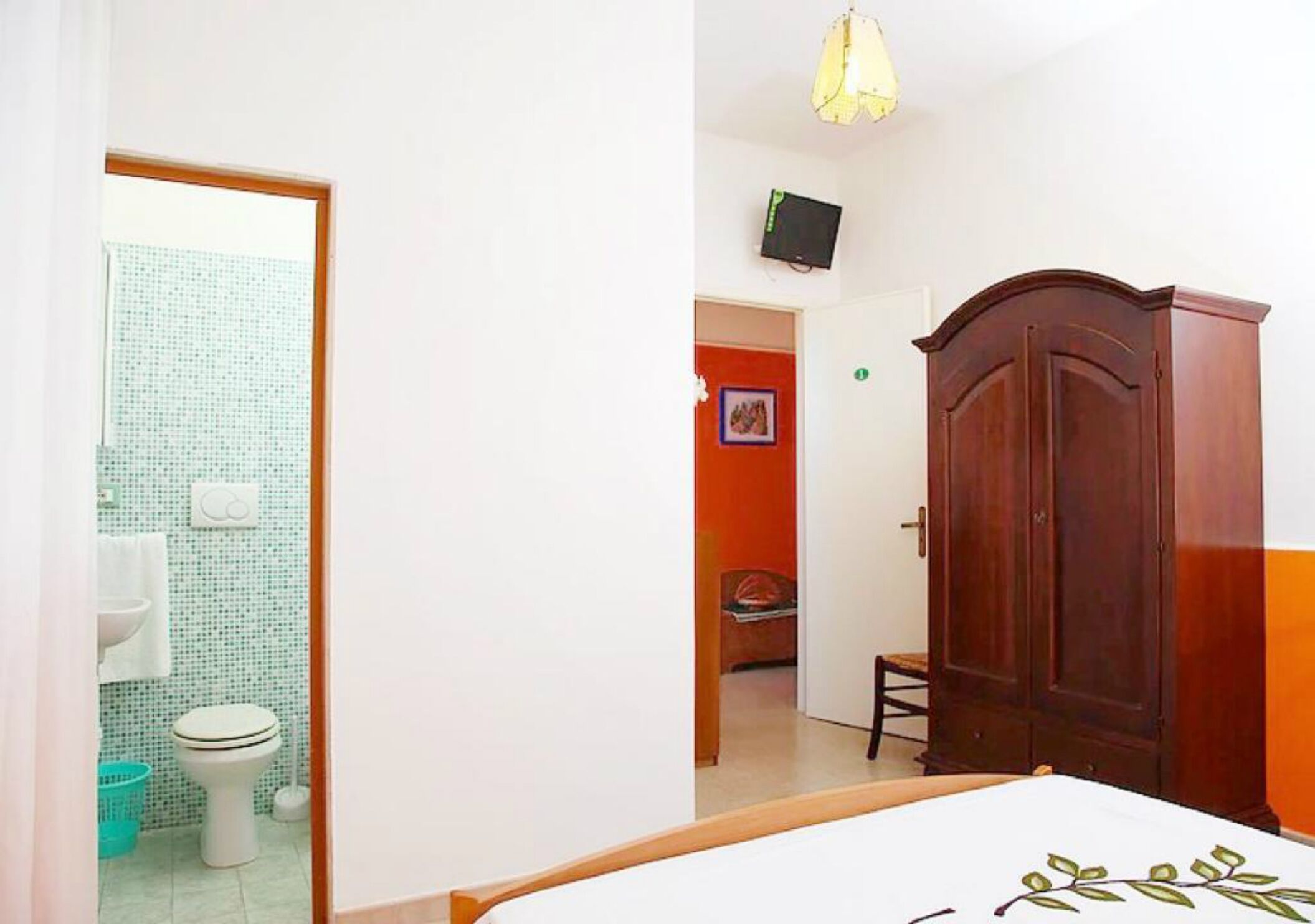 Een slaapkamer met een houten garderobe, een bed, een aan de muur bevestigde televisie en een zichtbare badkamer door een open deur.