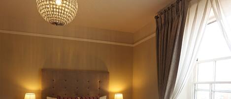 A hotel bedroom with a large bed, two bedside tables, a chandelier, and a window with curtains.