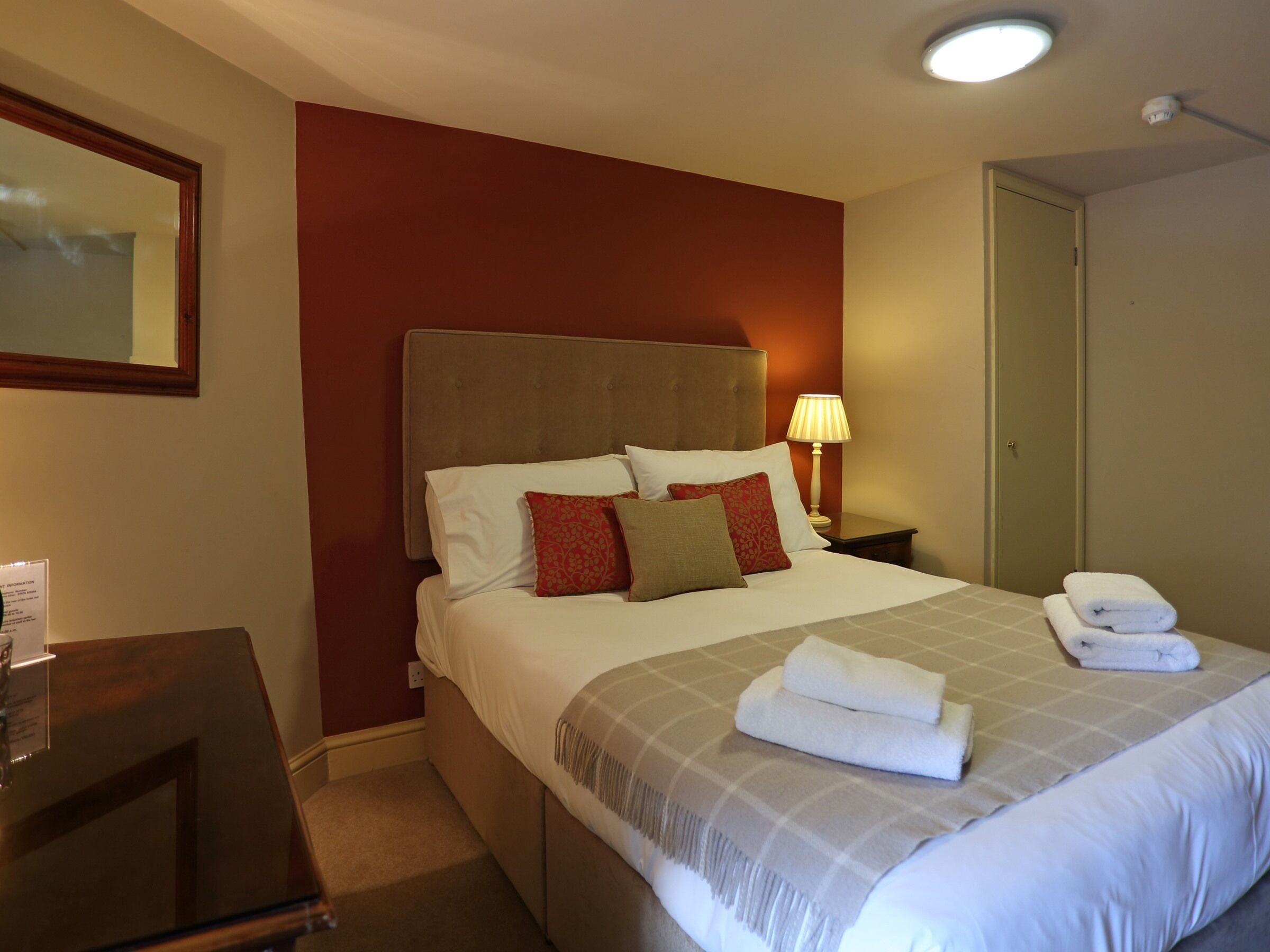 A hotel room with a large bed, a nightstand with a lamp, a mirror, and a door.