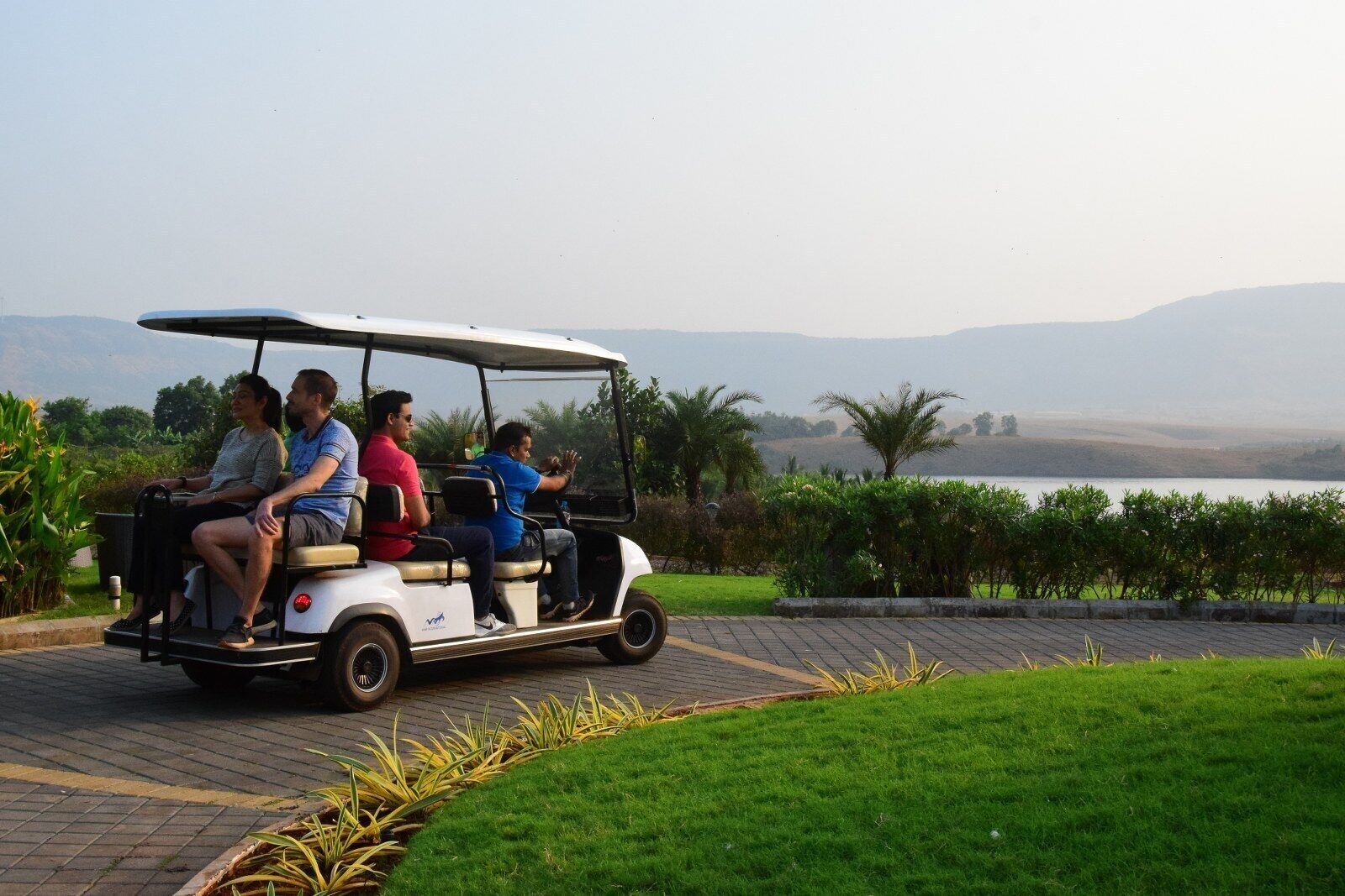 A golf cart with four passengers on a paved path with grass and plants on the sides.