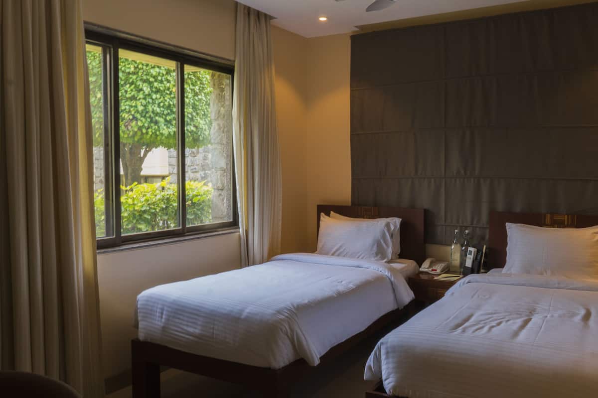 Une chambre d’hôtel avec deux lits simples, une table de chevet avec une lampe et une fenêtre avec des rideaux.