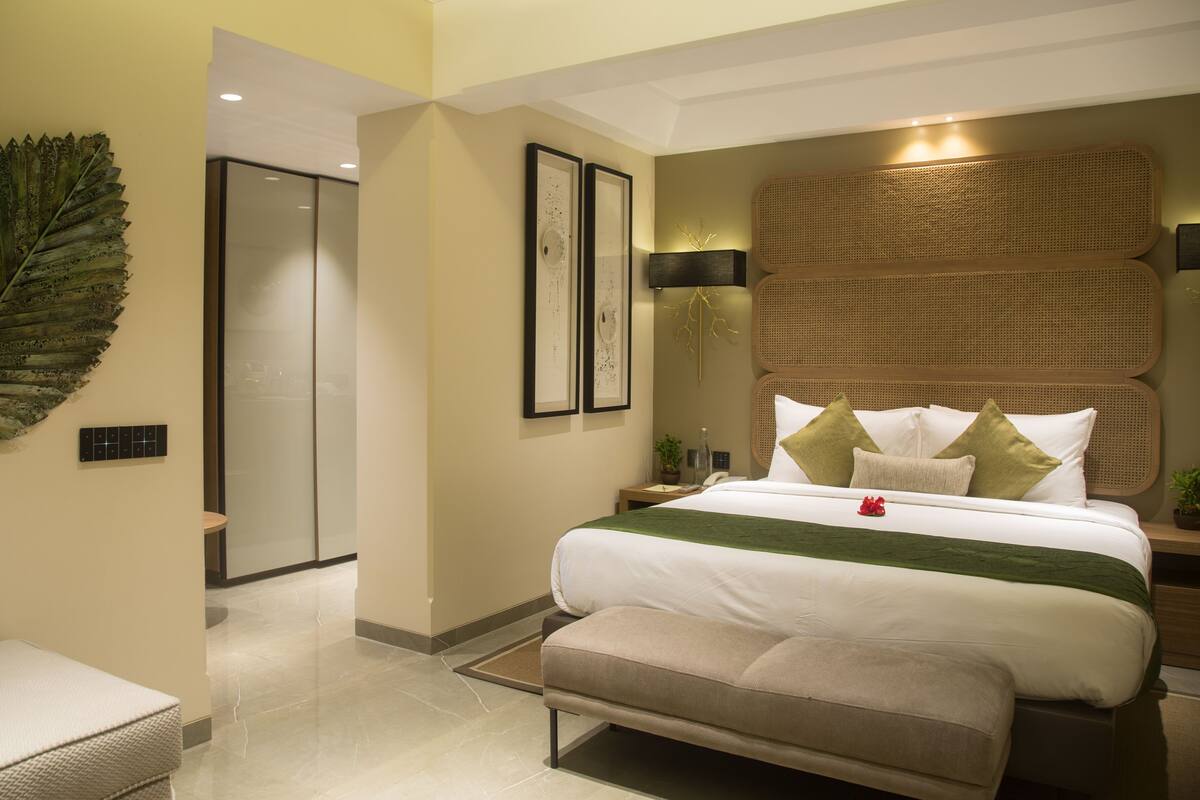 Une chambre d’hôtel moderne avec un grand lit, une tête de lit en rotin, un plaid vert et une grande décoration en forme de feuille sur le mur.