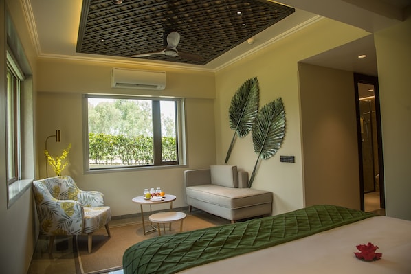 A hotel room with a bed, a sofa, a small table with drinks, a window with a view of greenery, and a ceiling fan.