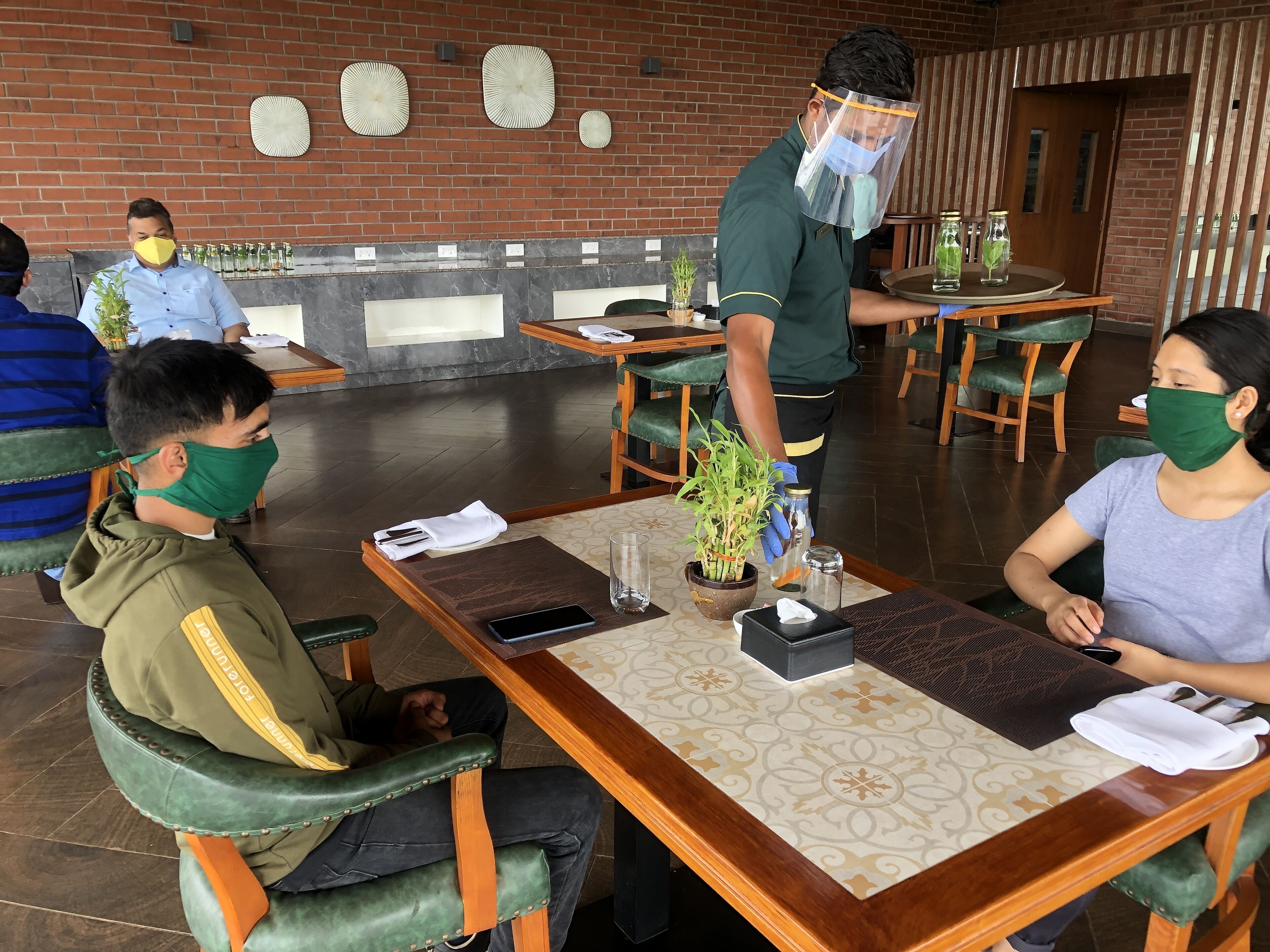 A restaurant with tables set for dining, a waiter attending to a table, and a brick wall with decorative plates in the background.