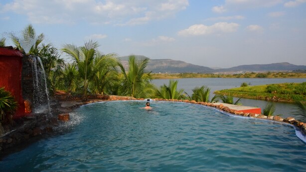 Una piscina con caída de agua, rodeada de palmeras y vista a colinas distantes.