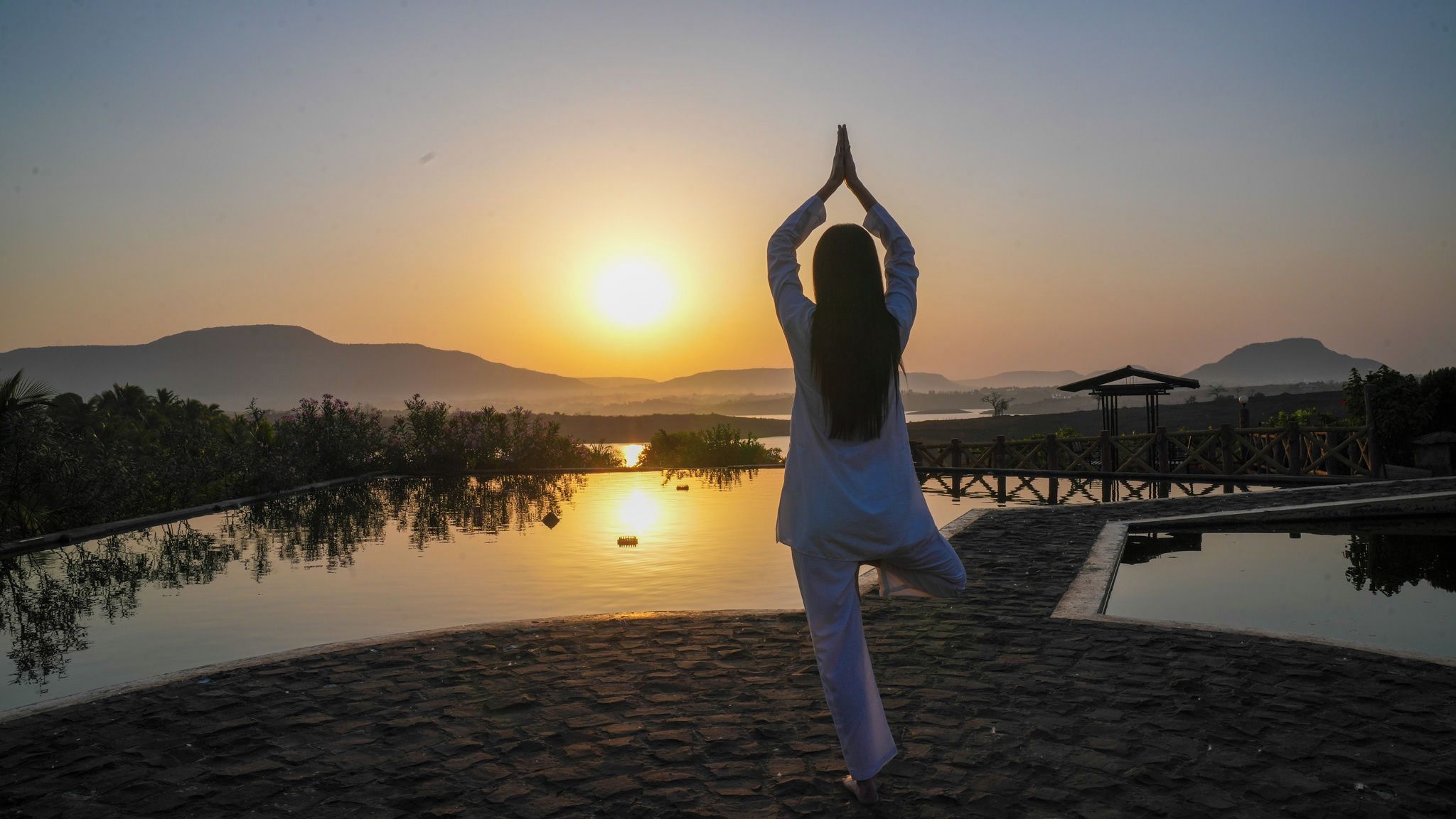 A person performing a yoga pose on a stone platform with a sunset view over a calm body of water and distant hills.