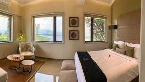 Un dormitorio con una cama grande, una silla, una mesita con fruta y dos ventanas con vistas a un lago y montañas.