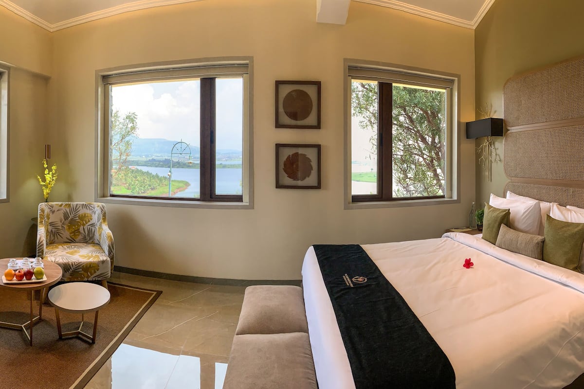 Une chambre avec un grand lit, une chaise, une petite table avec des fruits, et deux fenêtres donnant sur un lac et des montagnes.