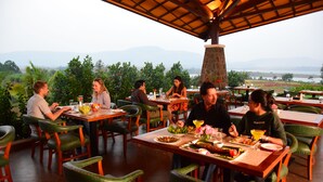 Un restaurante con terraza, varias mesas y sillas, rodeado de vegetación y vistas a montañas.