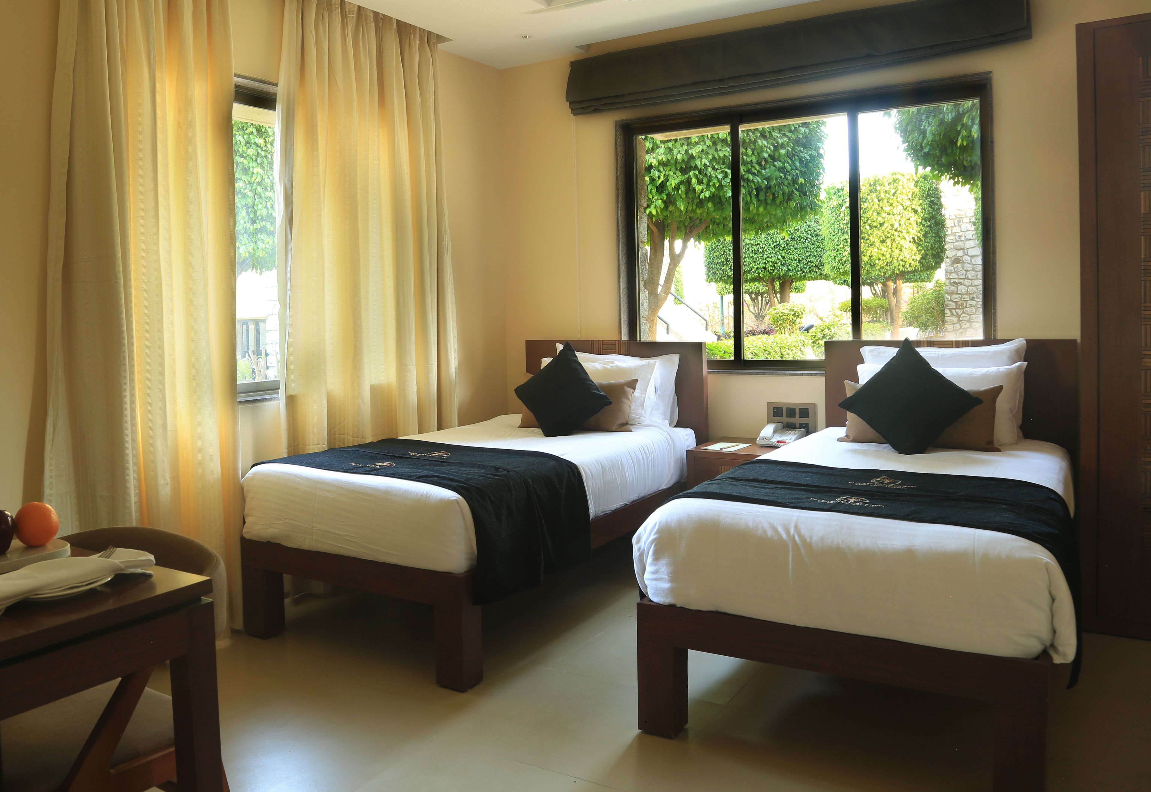 A hotel room with two single beds, a wooden desk, a telephone, and a window with a view of greenery.