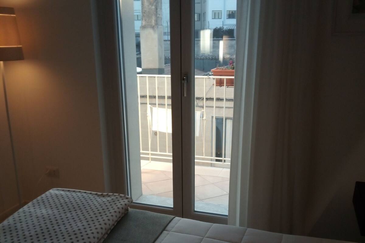 A hotel room with a bed, a window with a view of buildings, and a door.