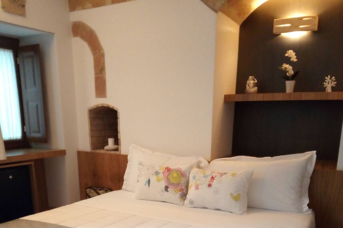 A neatly made bed with a grey duvet and white pillows, a wooden headboard, and a small shelf with decorative items.
