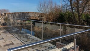 Un puente de piedra con barandilla de metal, un camino pavimentado y una masa de agua.