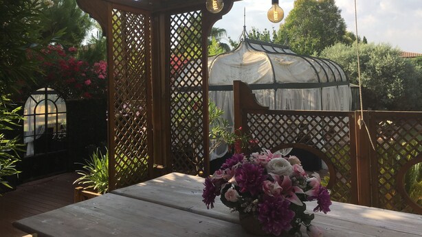 A wooden pergola with lattice panels, a table with a floral centerpiece, and a greenhouse in the background.