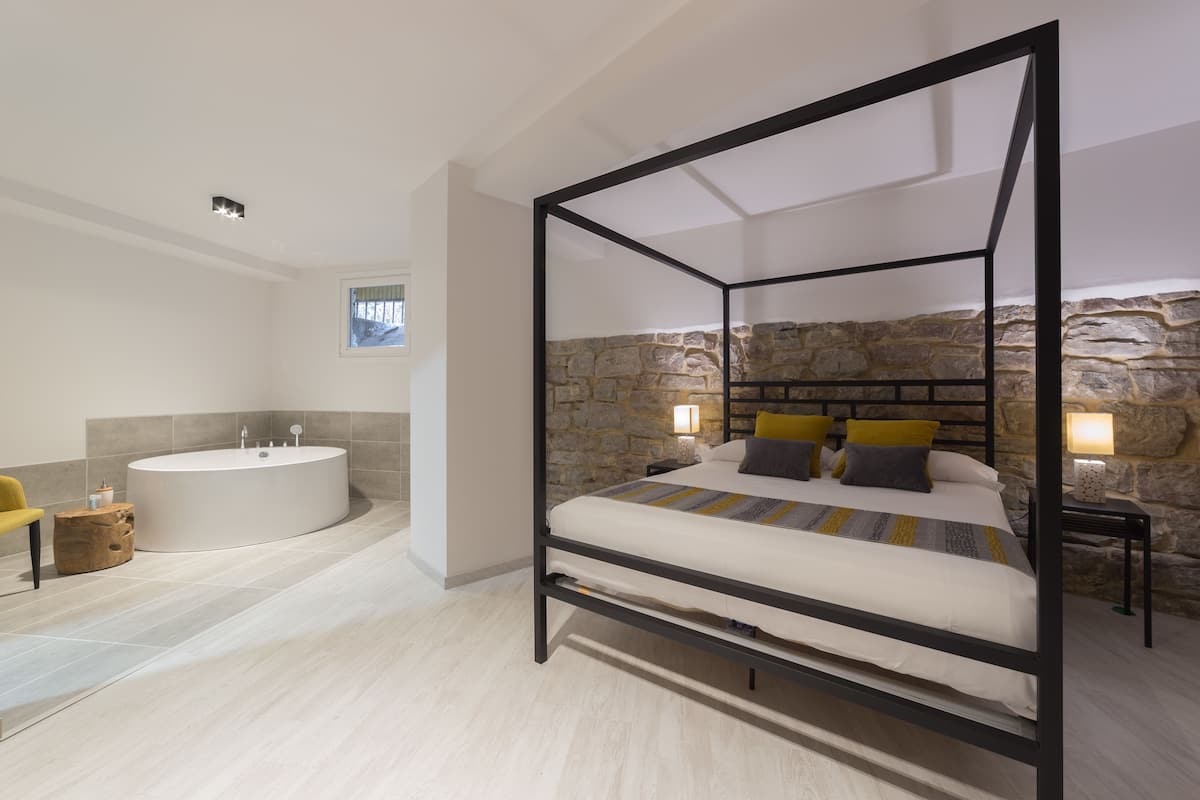 A modern bedroom with a four-poster bed, a large round bathtub, and a stone wall.
