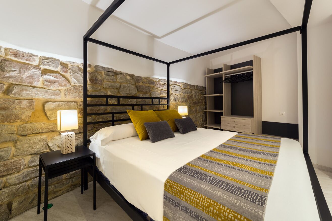 A bedroom with a stone wall, a four-poster bed, a nightstand with a lamp, and a built-in wardrobe.