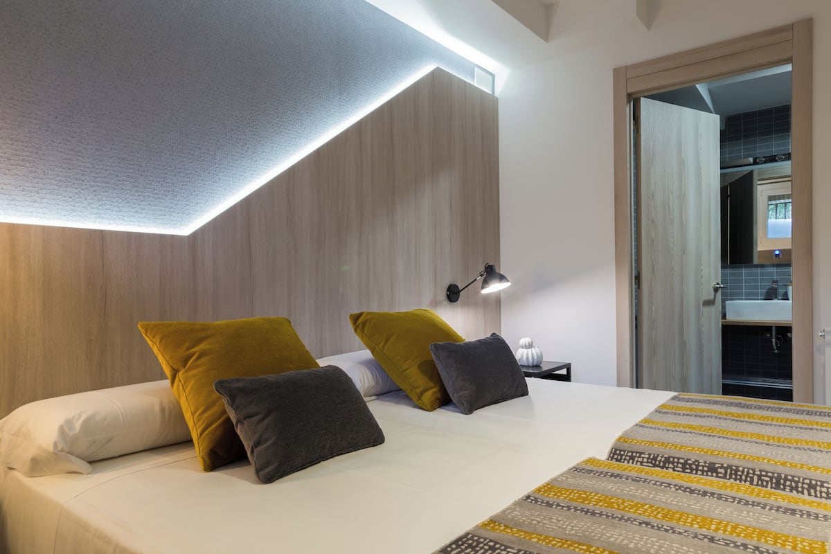 A modern hotel room with a bed, pillows, a bedside table, a lamp, and a view of a bathroom.