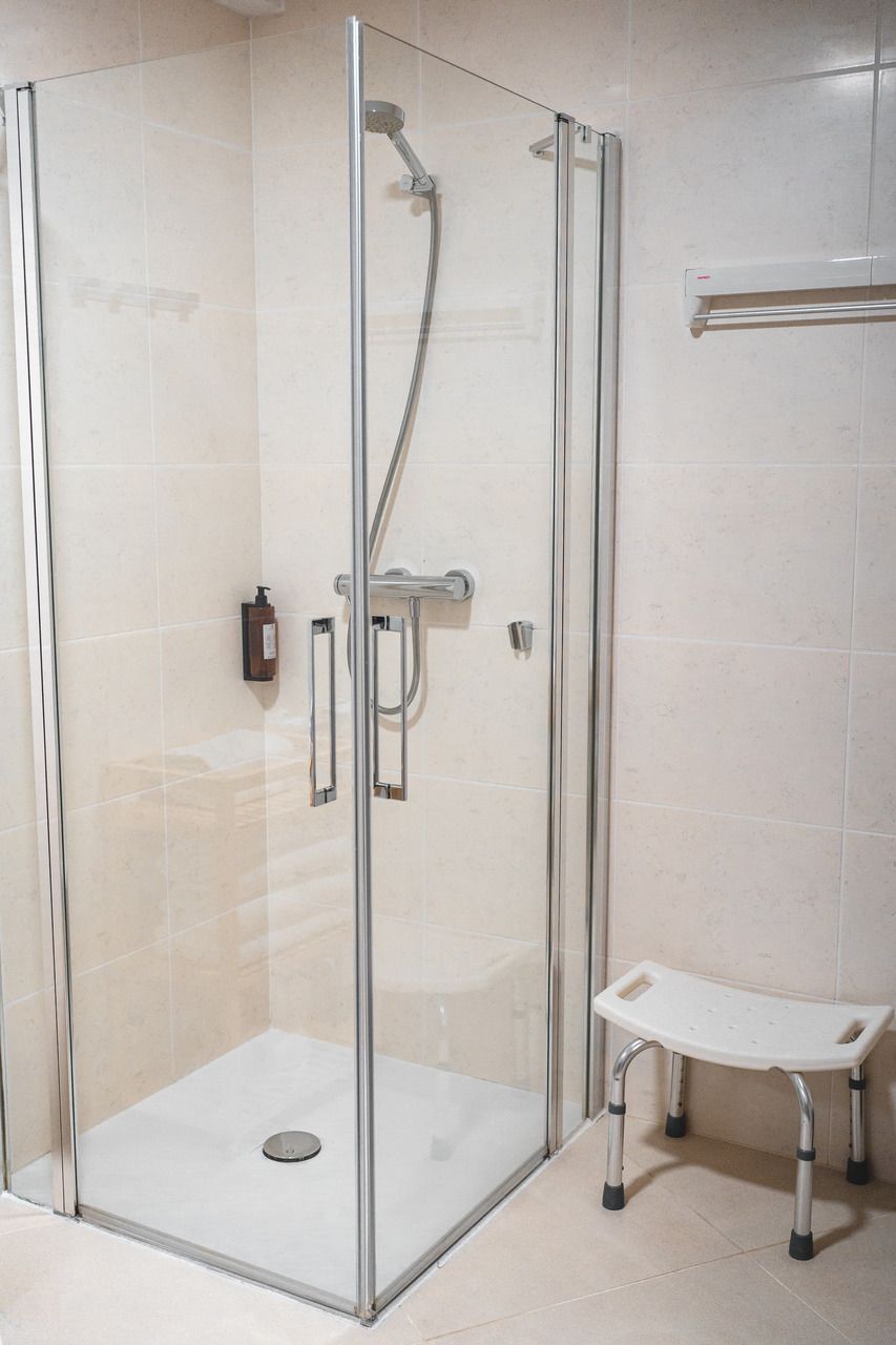 A glass-enclosed shower stall with a handheld showerhead, a handheld shower control, and a small bench with wheels.