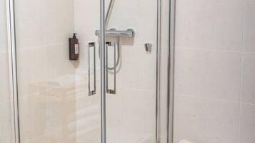 A glass-enclosed shower stall with a handheld showerhead, a handheld shower control, and a small bench with wheels.