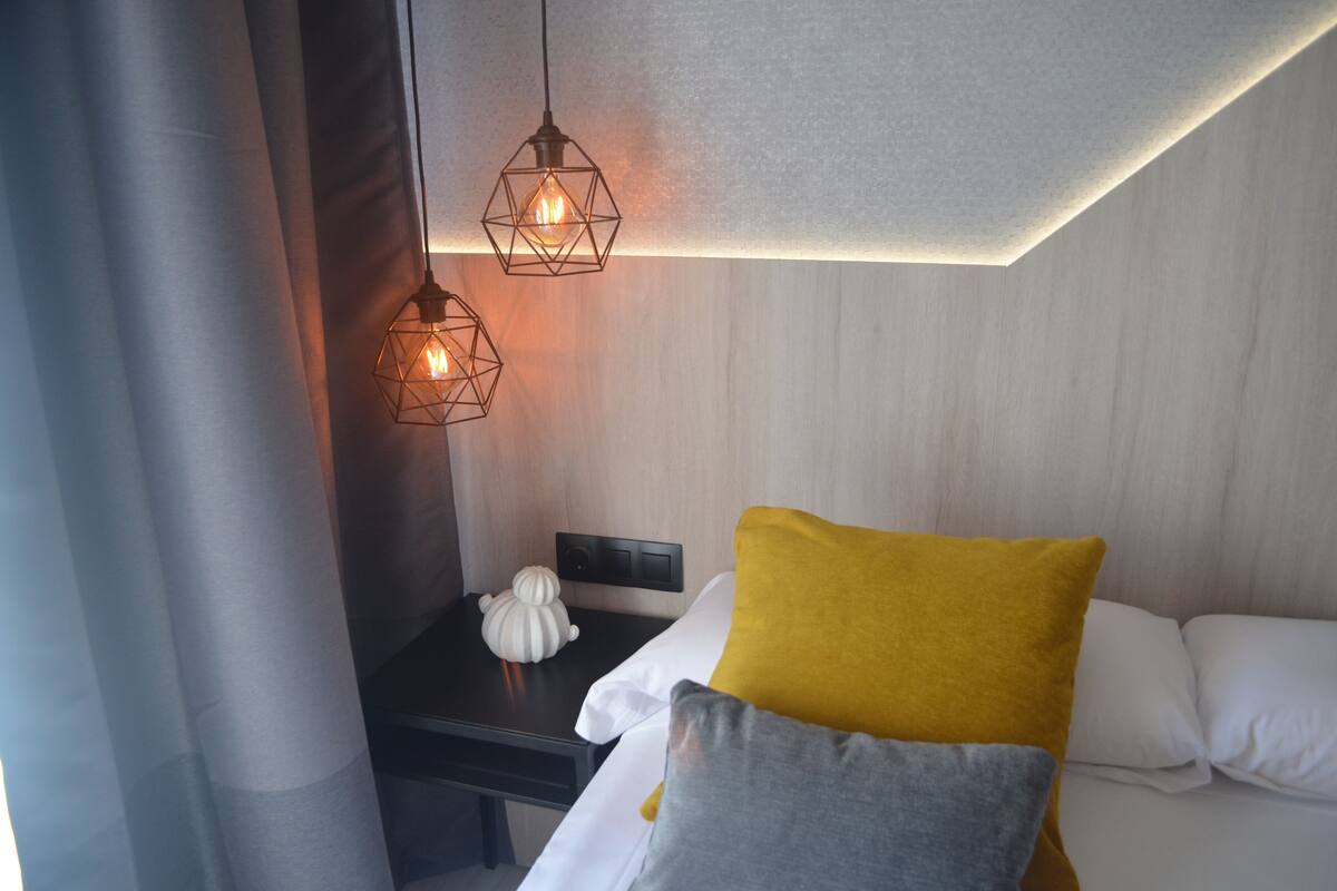 A hotel room with a bed, a bedside table, a wall-mounted control panel, and two geometric pendant lights.