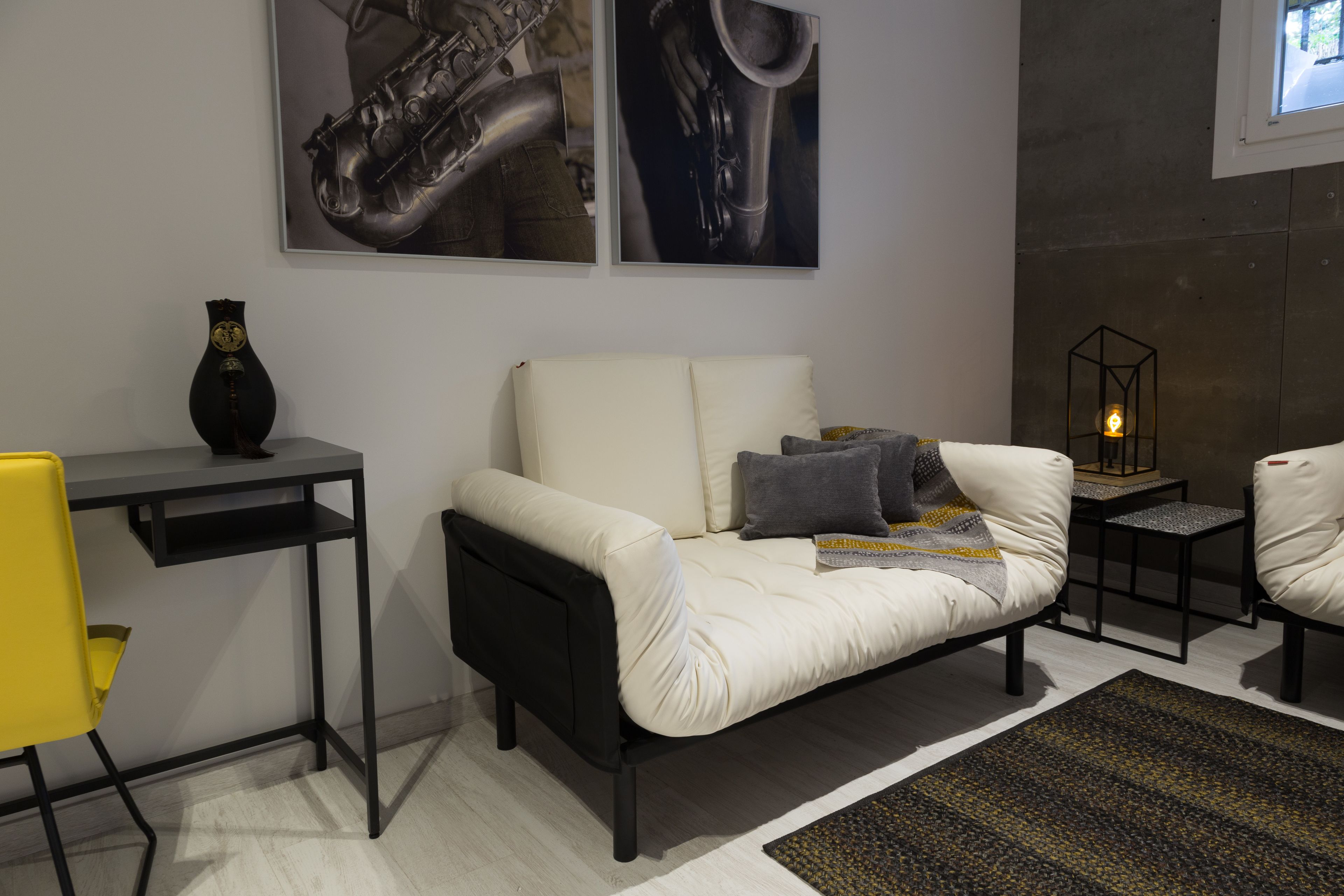 A modern living room with a white sofa, a small black table, and a yellow chair. There are two framed pictures on the wall and a decorative vase on the table.