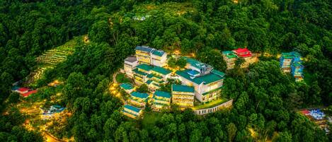 An aerial view of a resort with multiple buildings surrounded by dense forest, featuring terraced gardens and a winding road.