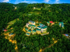 An aerial view of a resort with multiple buildings surrounded by dense forest, featuring terraced gardens and a winding road.