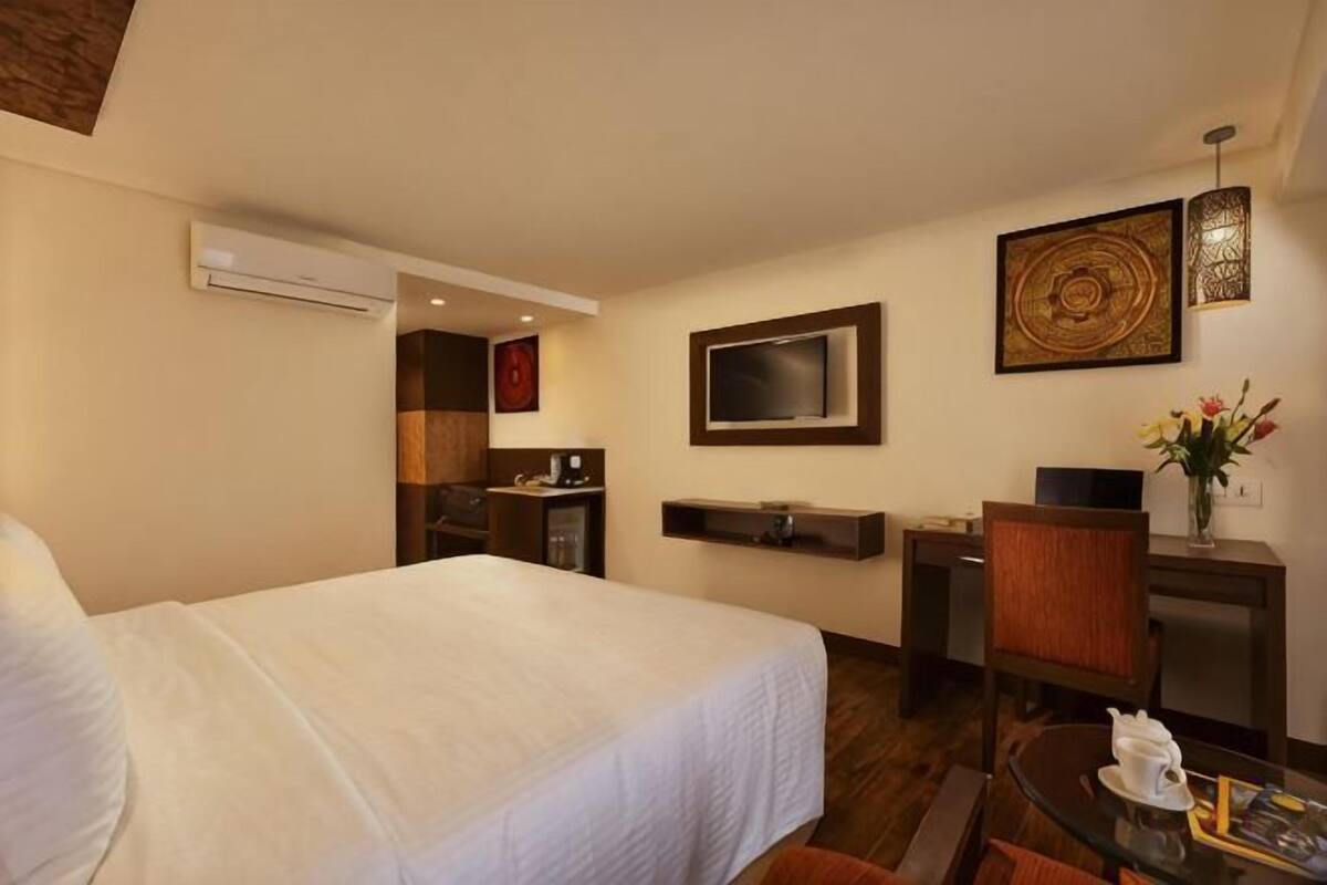Ein Hotelzimmer mit Bett, Schreibtisch, Stuhl, Fernseher, einer fest installierten Klimaanlage und einem kleinen Tisch mit einer Vase.