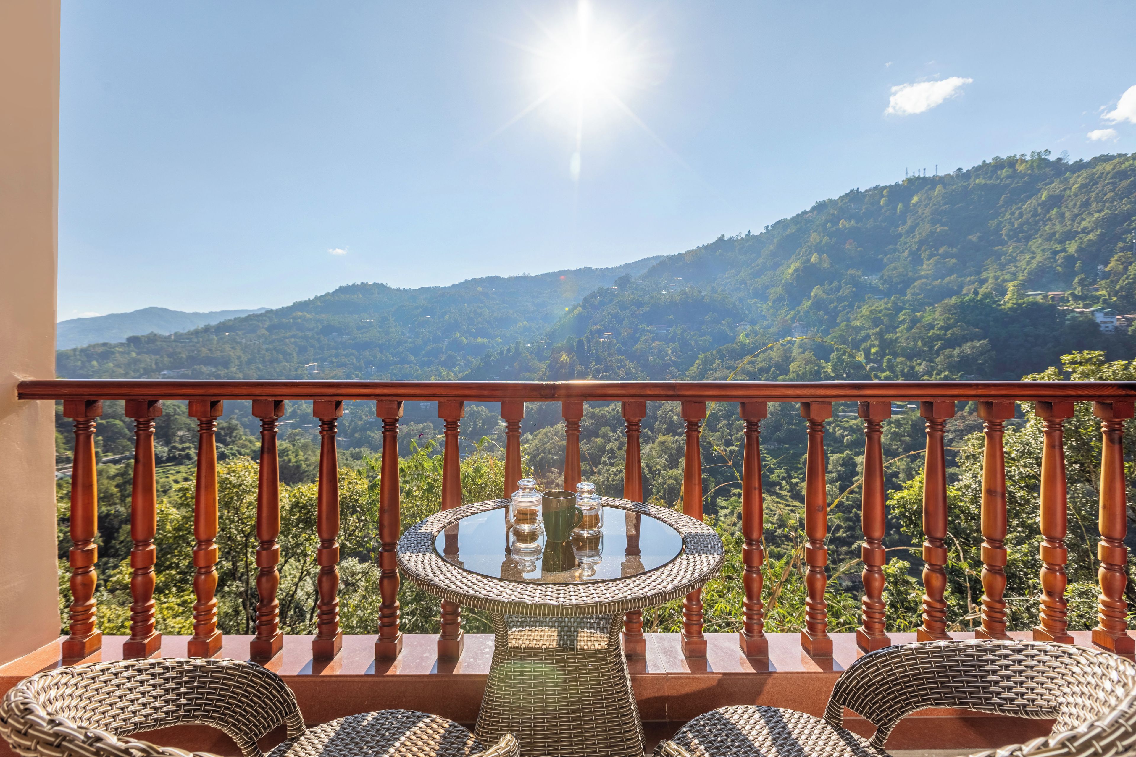 Ein Balkon mit einem runden Glastisch und Korbstühlen, mit Blick auf eine gebirgige Landschaft.