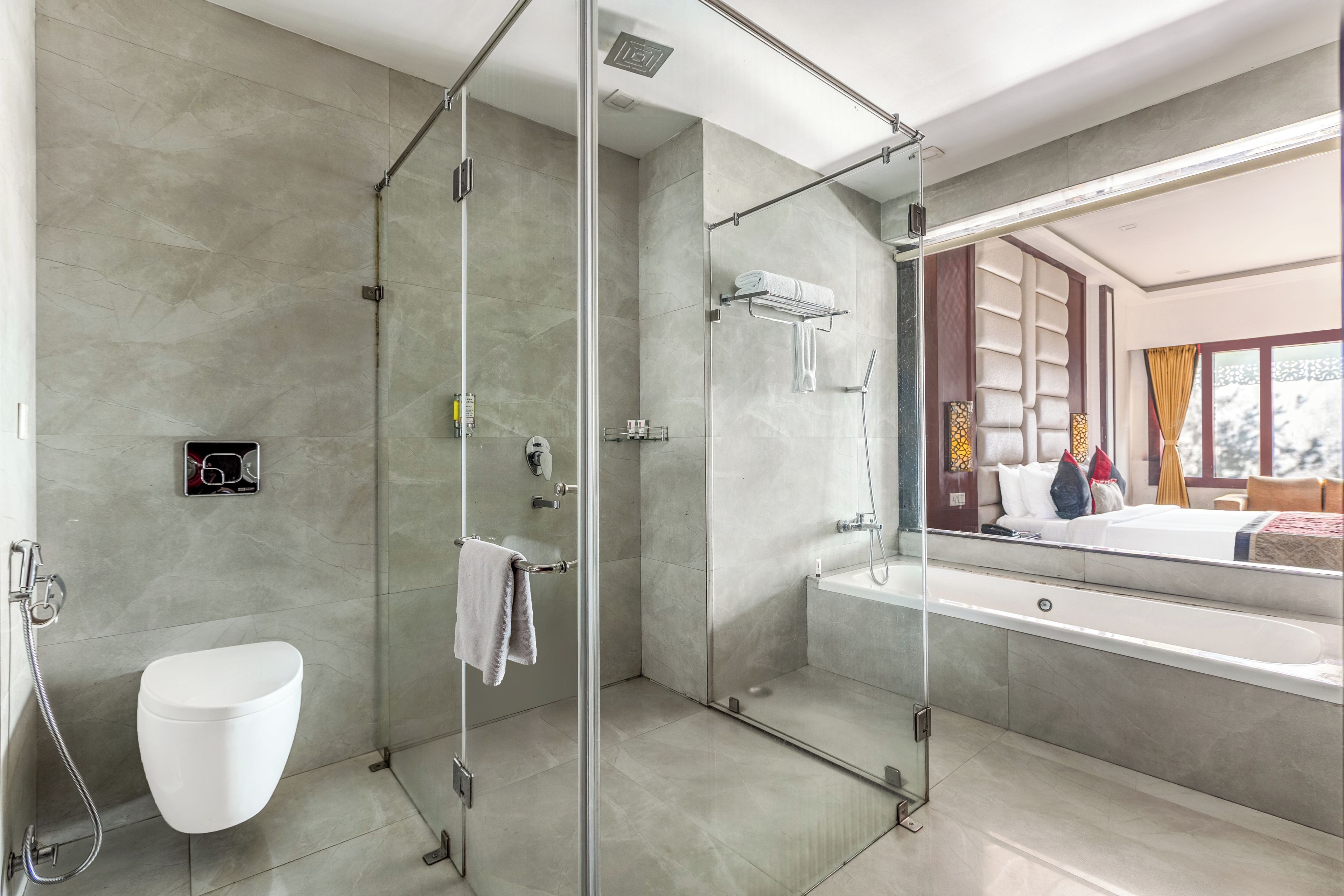 A modern bathroom with a glass-enclosed shower, a white toilet, and a large mirror reflecting a bedroom with a bed and window.