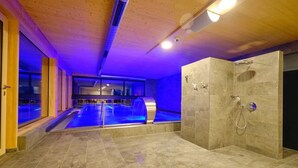 Piscina interior moderna con un elemento acuático iluminado en azul, zona de ducha separada y techo de madera.